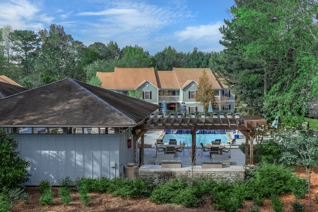 Outdoor swimming pool area with lounge chairs and seating under a pergola, with apartment buildings in the background.