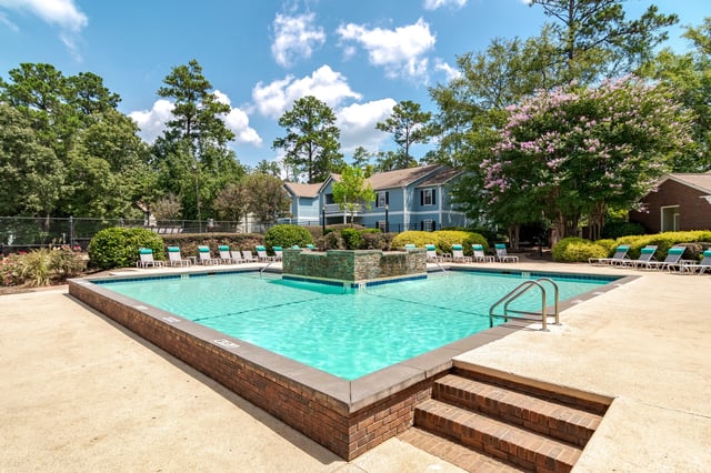 Swimming pool with lounge chairs and a water feature, surrounded by trees and apartment buildings.