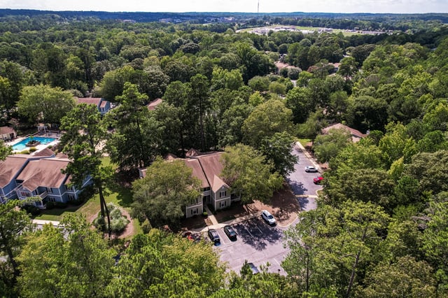 Aerial view of apartment buildings surrounded by lush green trees, with a swimming pool visible.