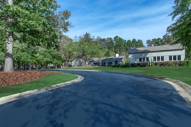 Curving asphalt road leading to a low-rise apartment building complex entrance with mature trees and manicured lawns.