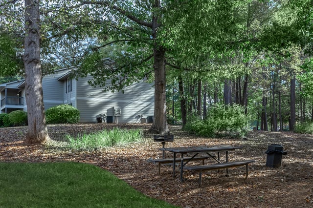 Picnic table and grill in a wooded area with apartment buildings in the background.