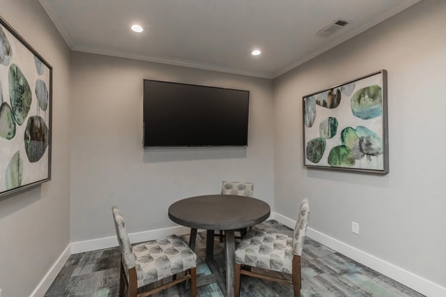 A table and chairs in a common area with a TV mounted on the wall and abstract art.
