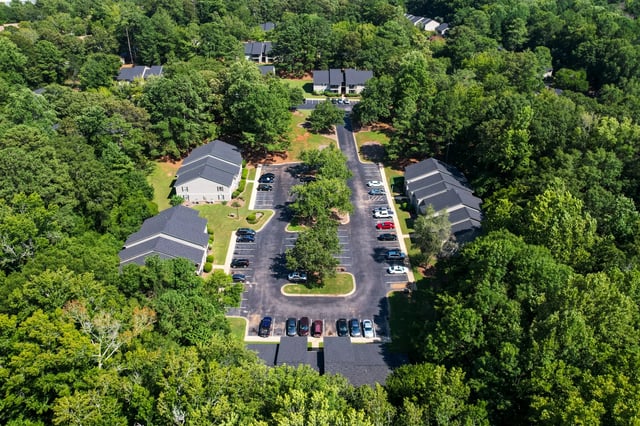 Aerial view of apartment buildings surrounded by lush green trees, with a parking lot in the center.