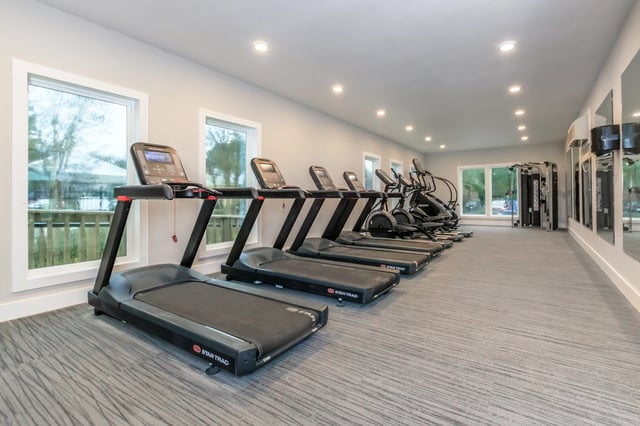Treadmills and elliptical machines in a well-lit fitness center with large windows.