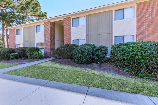Exterior view of apartment building with brick and green siding, manicured bushes, and a walkway.