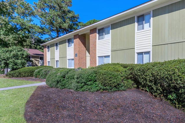 Exterior view of apartment building with green and white siding, brick accents, and manicured landscaping.