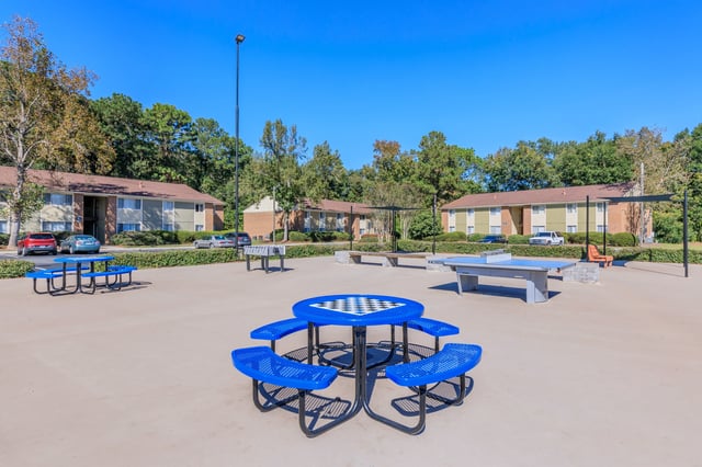Outdoor recreational area with chess table, ping pong table, and foosball table, with apartment buildings in the background.