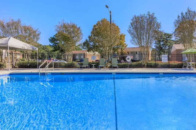Bright blue swimming pool with lounge chairs and umbrellas surrounding it. Buildings visible in the background.