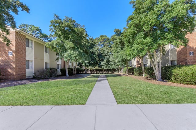 A paved walkway leads through a grassy courtyard lined with trees between apartment buildings.