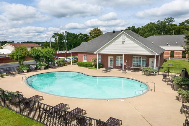 Resort-style swimming pool with lounge chairs and tables, adjacent to a brick clubhouse.