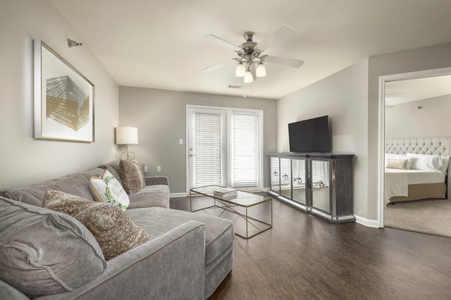 Spacious living room with a large sectional sofa, glass coffee table, and entertainment center with TV. A glimpse of the bedroom is visible through an open doorway.