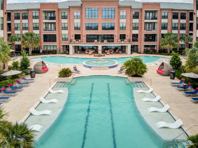 Modern apartment complex with a large swimming pool and hot tub area, surrounded by lounge chairs and palm trees.