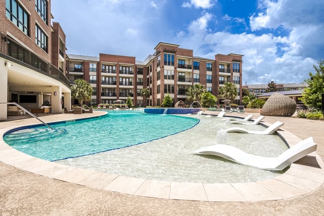 Outdoor swimming pool with lounge chairs and fountains, adjacent to a brick apartment building.
