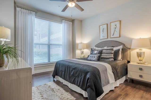 A spacious bedroom featuring a king-sized bed with plush bedding, a ceiling fan, and large windows with white curtains.
