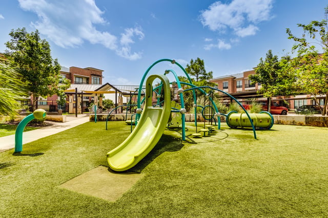 A brightly lit playground with a green slide and tunnel, artificial turf, and apartment buildings in the background.