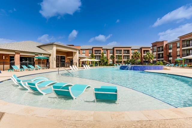 Resort-style swimming pool with in-water lounge chairs and a spa area, surrounded by apartment buildings.