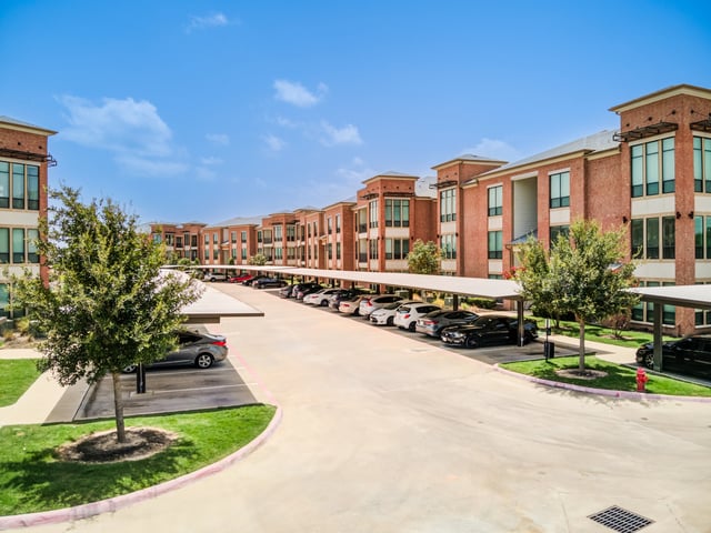 Covered parking lot with cars parked, alongside brick apartment buildings under a blue sky.