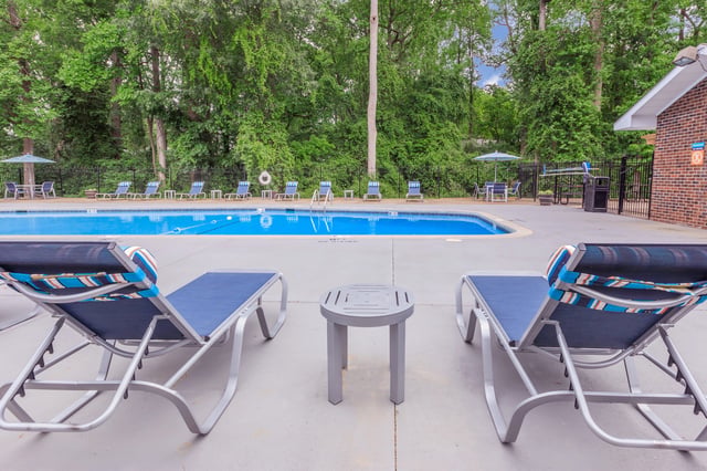 Poolside lounge chairs and a table in front of a swimming pool with surrounding trees.