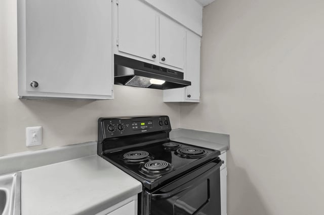 Black electric stove with four burners and oven, beneath white cabinets and a black range hood.
