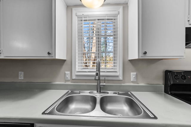 Kitchen sink with stainless steel double basin and faucet, window with blinds in the background.