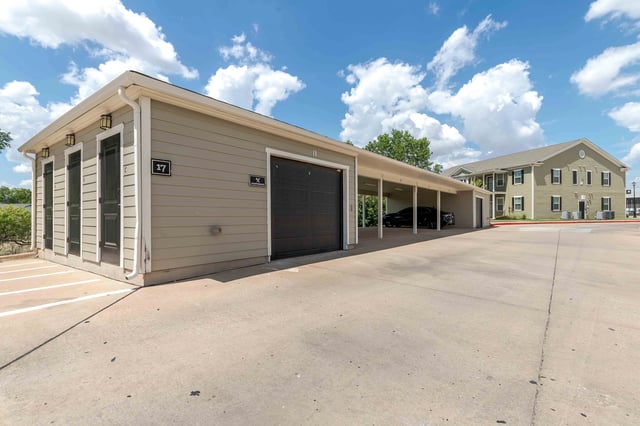 Exterior view of a garage and carport with apartments in the background.