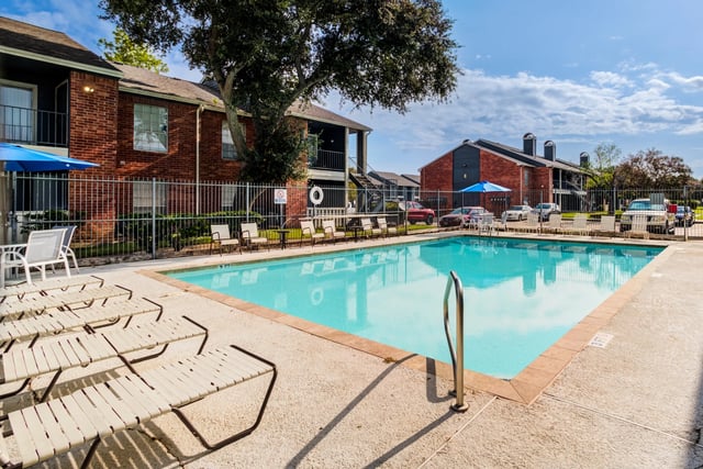 Sparkling swimming pool with lounge chairs and umbrellas at a brick apartment complex.