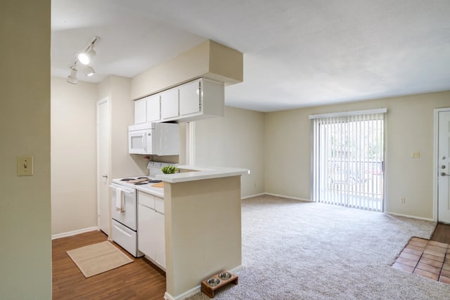Kitchenette with white cabinets and appliances, open to a living area with carpet and sliding glass doors.