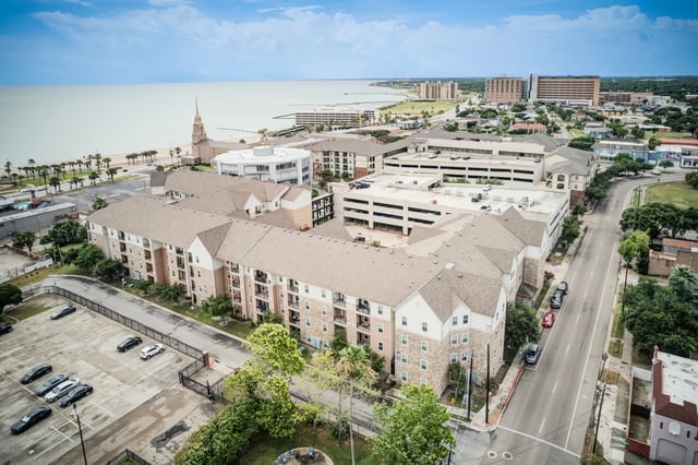 Aerial view of apartment buildings near the ocean with a parking lot and street in the foreground.
