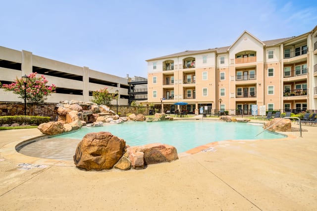 Resort-style swimming pool with a rock waterfall and lounge chairs in front of a tan apartment building.