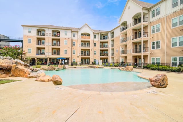 Resort-style swimming pool with a rock waterfall feature and lounge chairs, adjacent to a modern apartment building.