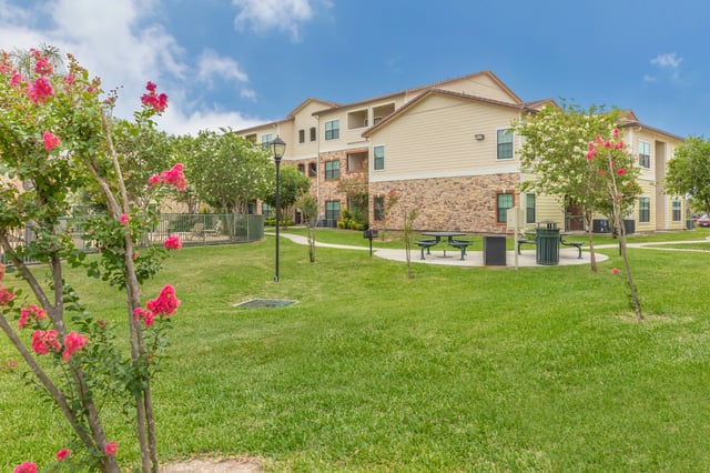 Exterior view of apartment buildings with lush green lawns, picnic tables, and flowering trees.