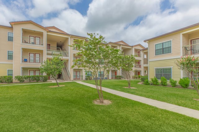 Exterior view of apartment buildings with balconies and walkways, surrounded by green grass and trees.