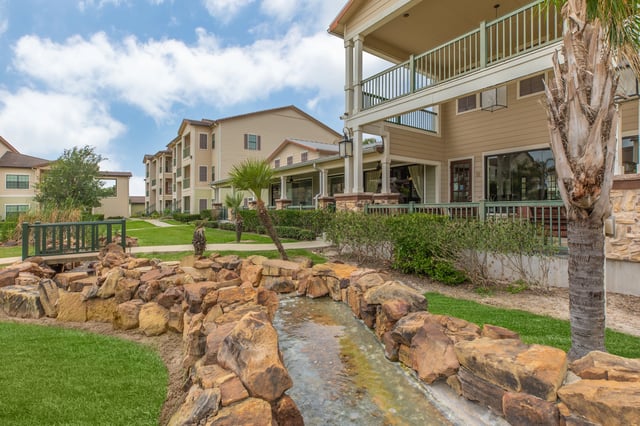 Outdoor decorative water feature with rocks and a small bridge in front of apartment buildings.