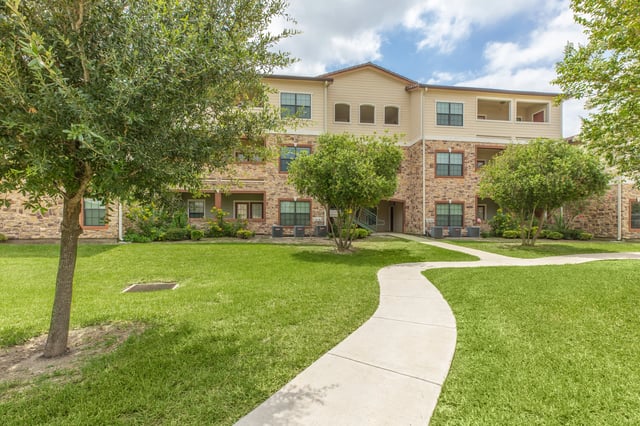 Exterior view of apartment building with stone and beige siding, surrounded by green grass and trees.