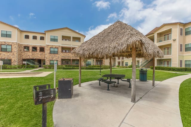 Outdoor picnic area with a thatched roof pavilion, picnic table, grill, and trash can, with apartment buildings in the background.