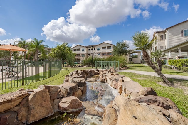 Outdoor water feature with rocks and a small bridge, with apartment buildings in the background.