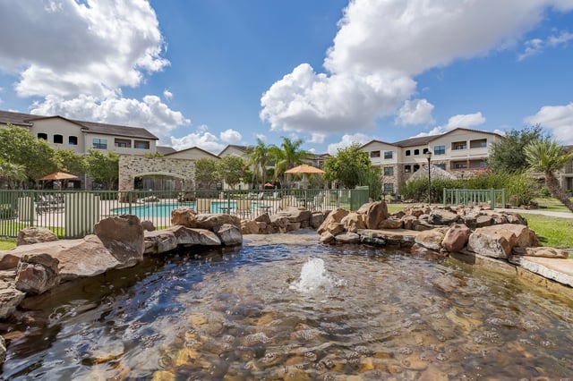 A serene water feature with rocks and a fountain in the foreground, with a swimming pool and apartment buildings in the background.