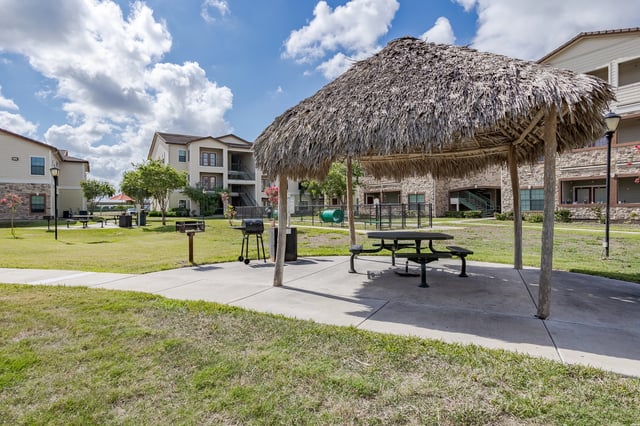 Outdoor picnic area with thatched roof, picnic tables, and grills with apartment buildings in the background.