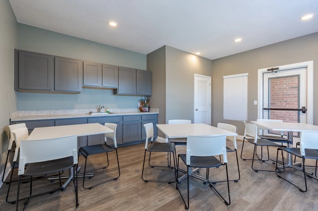 Community room with tables and chairs, featuring a kitchenette area.