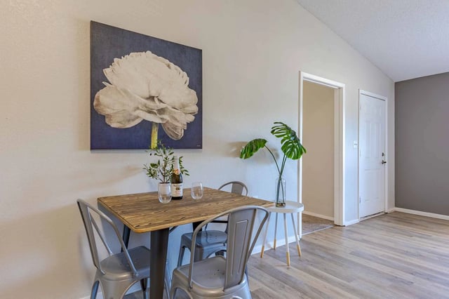 Dining area with a small wooden table, four metal chairs, a large floral painting, and a bottle of wine.