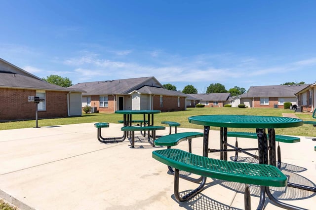 Picnic tables and grills are set up on a concrete patio area outside apartment buildings.