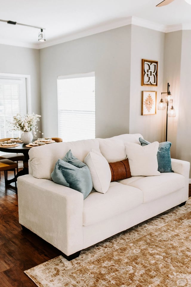 Comfortable living room with a white sofa, decorative pillows, and a dining table in the background.