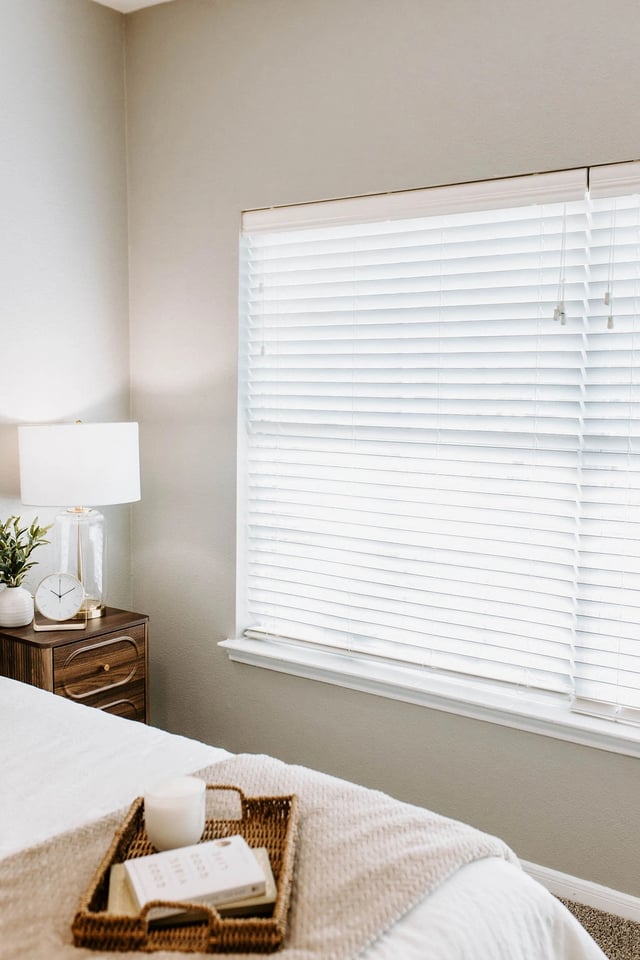 Bedroom with white blinds covering a large window, a nightstand with a lamp and clock, and a bed with a tray holding a book and candle.