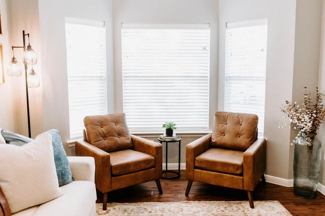 Two brown leather accent chairs flank a bay window with blinds, in a living room setting.