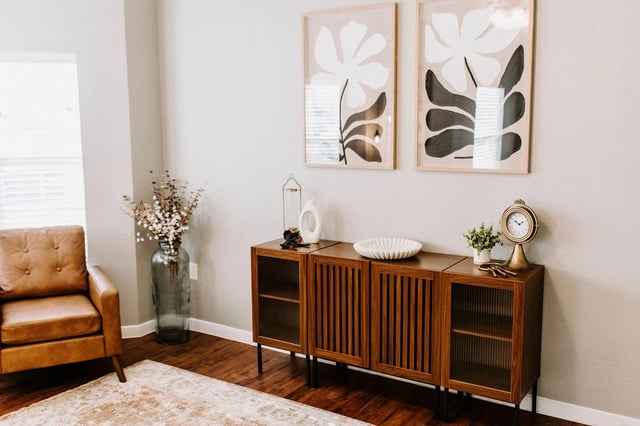 Modern living room with a brown leather armchair, a large vase of dried flowers, a wooden credenza with artwork above, and a decorative clock.