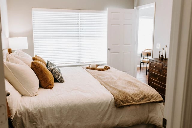 A well-made bed with plush pillows and a textured throw blanket, next to a window with blinds.