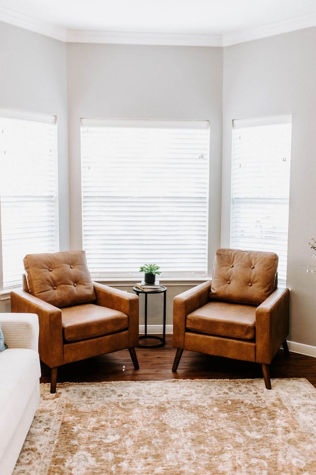 Two brown leather accent chairs in a living room with large windows.