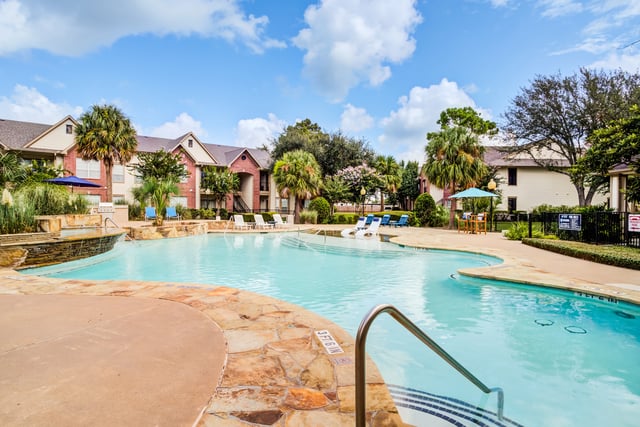 Large outdoor swimming pool with lounge chairs and umbrellas, adjacent to apartment buildings.
