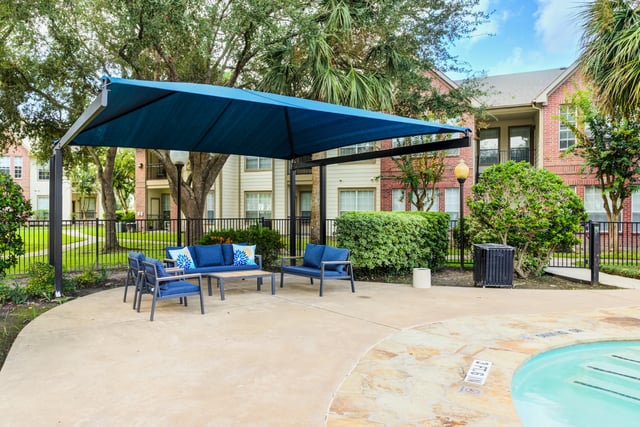 Outdoor seating area with blue couches and chairs under a blue canopy, next to a swimming pool.