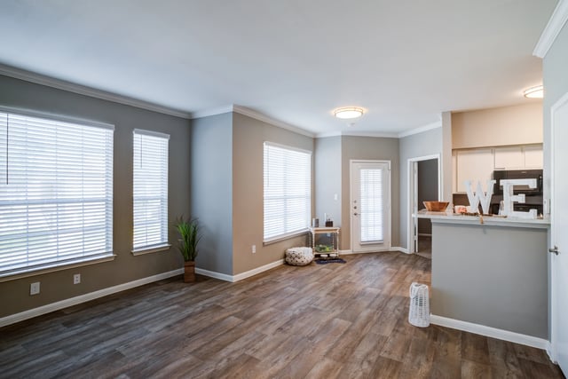 Living room and kitchen area of an apartment with large windows and hardwood floors.